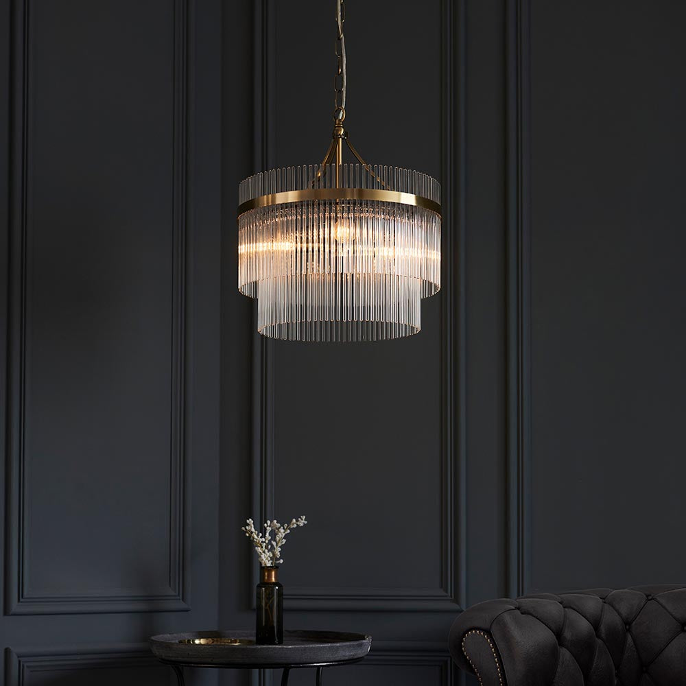 Endon Beaumont 3 Light Pendant in brass with two tiers of clear glass rods, illuminated above a small side table against a dark panelled wall.