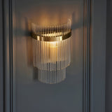 Beaumont Wall Light in brass, tiered clear glass rods surrounding a lit bulb on a round brass backplate, mounted on a grey panelled wall.