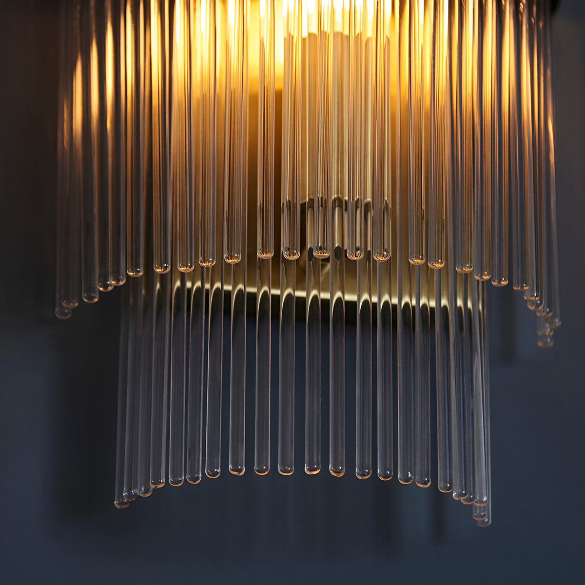 Beaumont Wall Light in brass, close-up of clear glass rods cascading from a brass backplate, glowing warmly against a dark background.