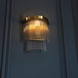 Beaumont Wall Light in brass, tiered clear glass rods diffusing warm light from a compact brass backplate, mounted on a dark panelled wall.
