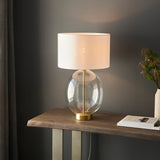 Brockmere Table Lamp in brass with an oversized oval clear glass base and white fabric drum shade, shown lit on a wooden console table.