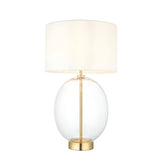 Brockmere Table Lamp with an oversized oval clear glass base, satin brass stem and base plate, and a white fabric drum shade on a plain background.