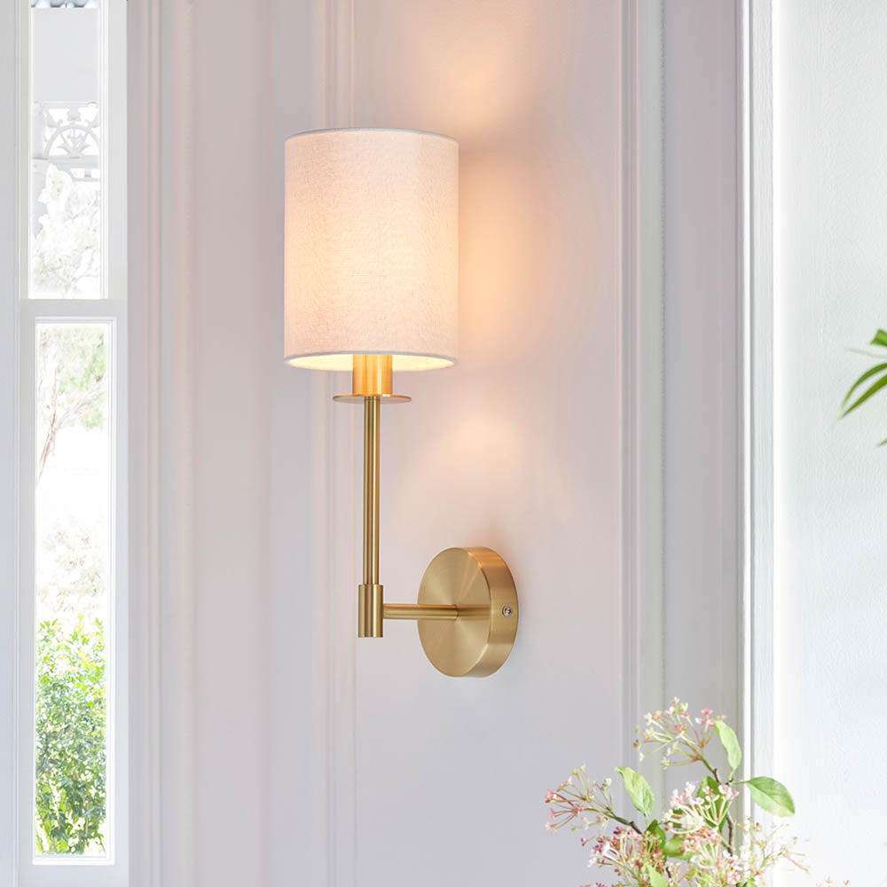 Endon Cliffhurst wall light with a satin brass upright and vintage white cylinder shade, illuminated on a panelled hallway wall beside a bright window.