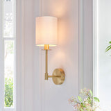 Endon Cliffhurst wall light with a satin brass upright and vintage white cylinder shade, illuminated on a panelled hallway wall beside a bright window.