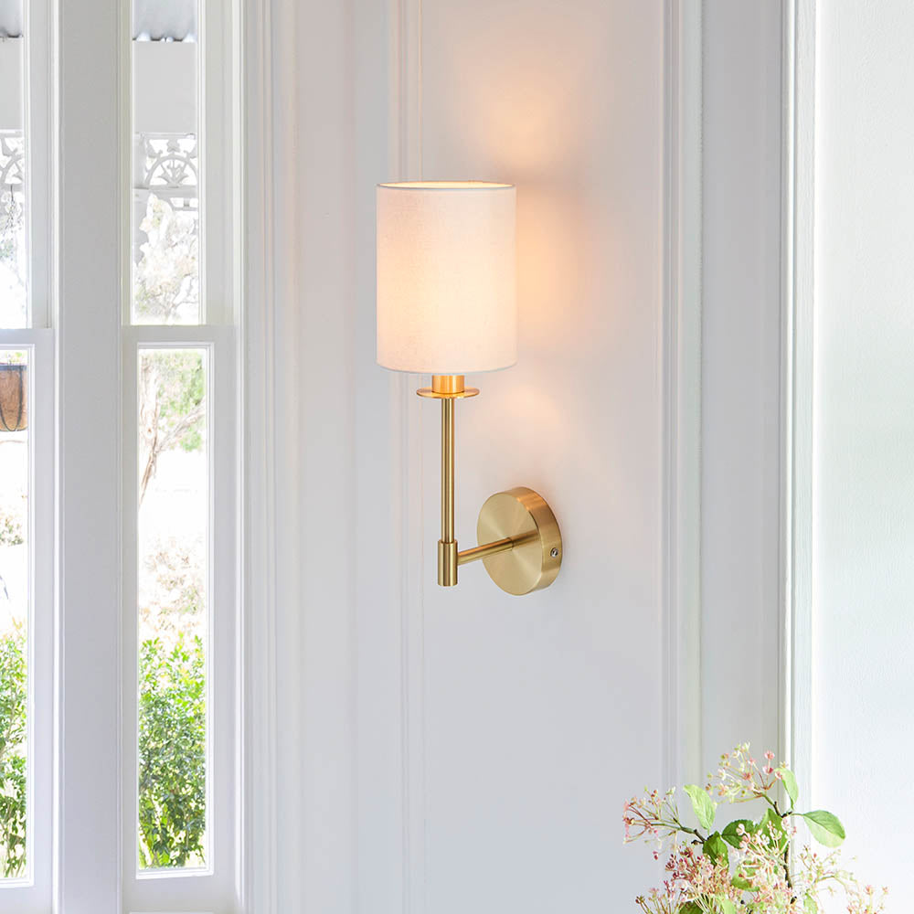 Endon Cliffhurst wall light in satin brass with a slim upright, round backplate and vintage white cylinder shade, mounted on a bright white panelled wall.