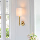 Endon Cliffhurst wall light in satin brass with a slim upright, round backplate and vintage white cylinder shade, mounted on a bright white panelled wall.