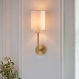 Endon Cliffhurst wall light in satin brass with a slim upright and vintage white cylinder shade, illuminated on a panelled wall in a living space.