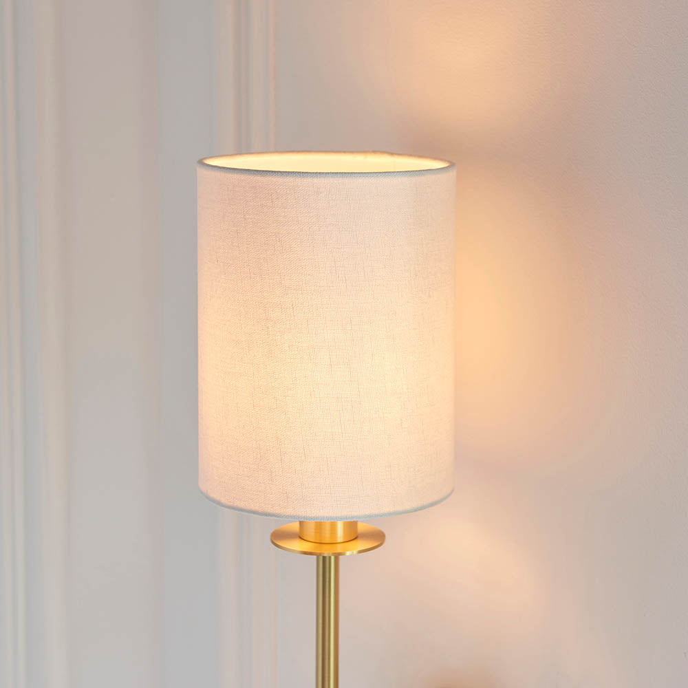 Endon Cliffhurst wall light with a slim satin brass upright and vintage white cylinder shade, illuminated against a pale panelled wall.