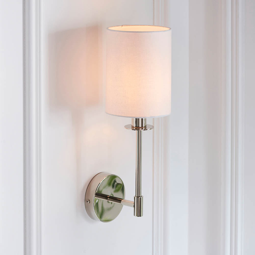 Endon Cliffhurst wall light with a slim polished nickel upright, round backplate and white cylinder fabric shade, mounted on a white panelled wall.