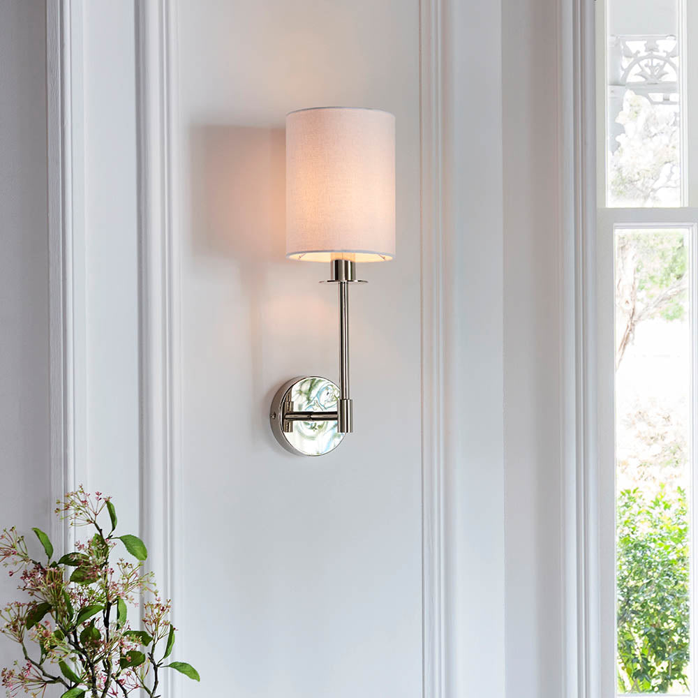 Endon Cliffhurst wall light in polished nickel with a slim upright, round backplate and vintage white cylinder shade, mounted on a white panelled wall.