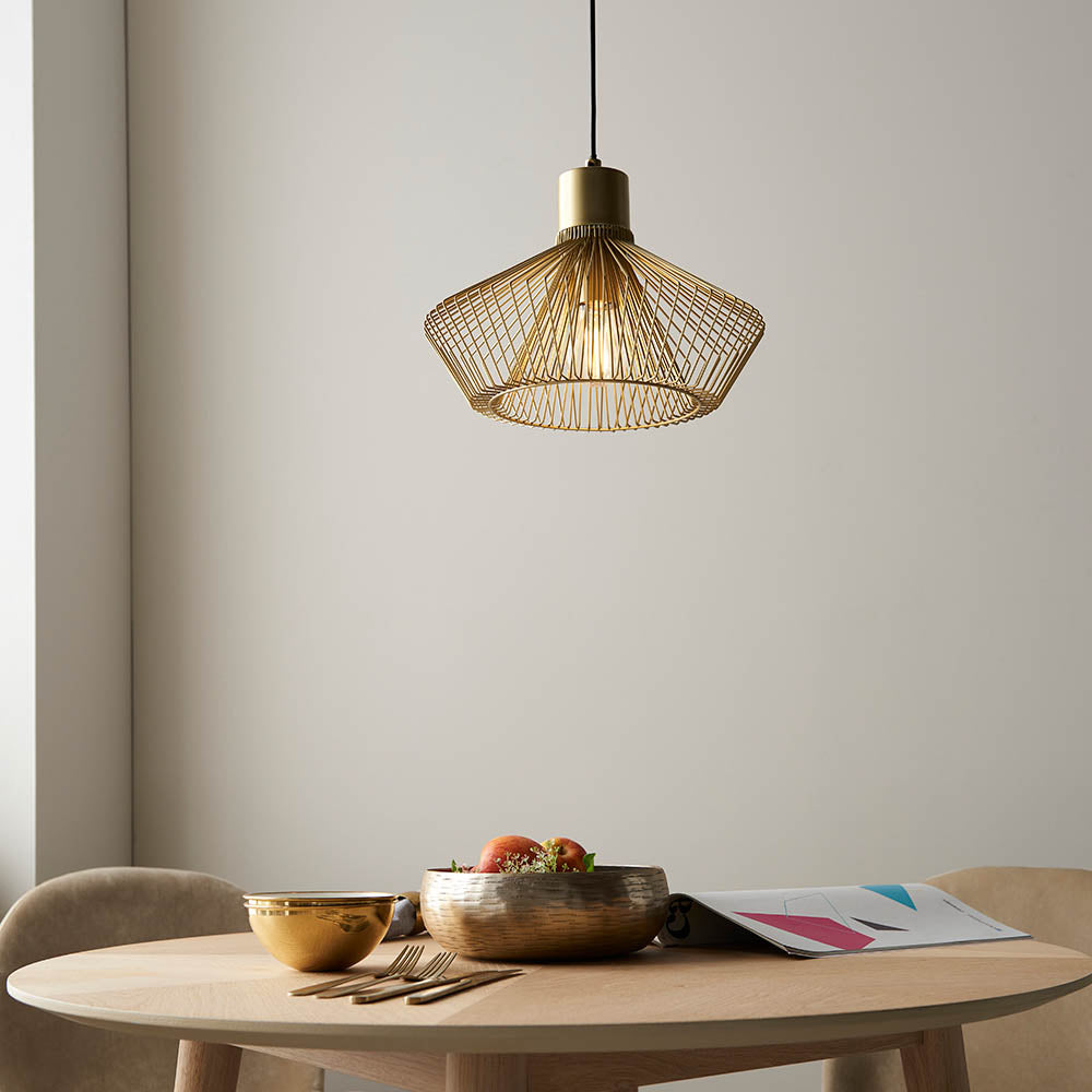 Endon Brockton gold cage pendant light with open metal shade and black flex, hanging above a wooden dining table set with bowls and a magazine.