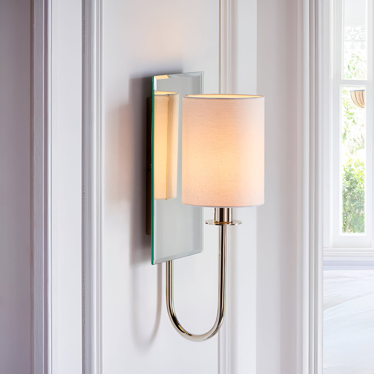 Endon Carlhurst wall light with bevelled mirrored backplate, polished nickel curved arm and vintage white fabric cylinder shade, mounted between windows.
