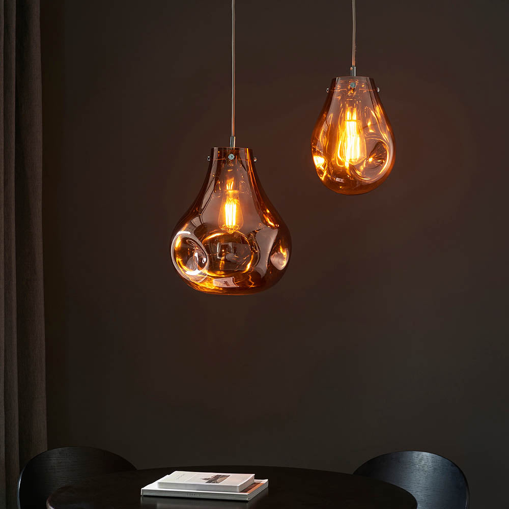 Two Brackwell copper glass pendant lights with organic molten shapes and chrome fittings, hanging above a black dining table.