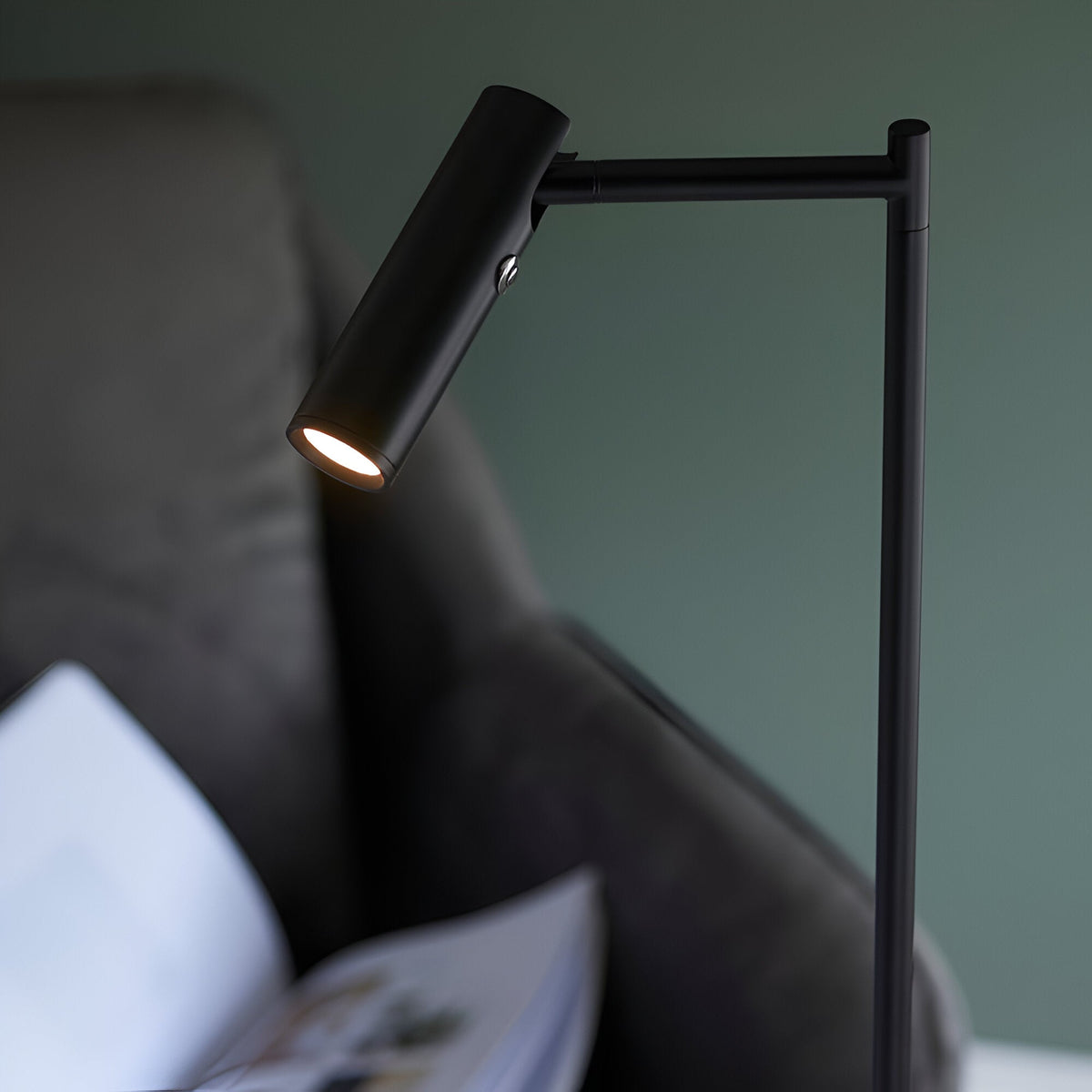 Endon Brackbourne floor lamp in matt black with an adjustable cylindrical LED head and push button switch, angled over a dark sofa in a bedroom setting.