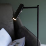 Endon Brackbourne floor lamp in matt black with an adjustable cylindrical LED head and push button switch, angled over a dark sofa in a bedroom setting.