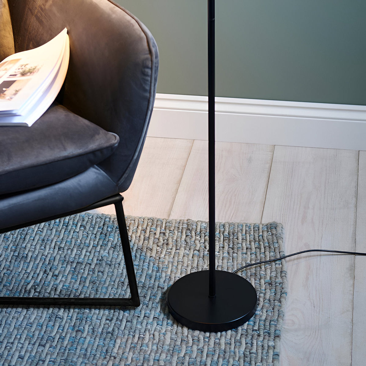 Endon Brackbourne floor lamp in matt black with round base and slim upright, set on a woven grey rug beside a dark velvet chair on pale wood flooring.