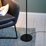 Endon Brackbourne floor lamp in matt black with round base and slim upright, set on a woven grey rug beside a dark velvet chair on pale wood flooring.