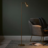 Endon Brackbourne brass LED floor lamp with adjustable cylindrical head beside a grey velvet armchair on a woven rug against a dark green wall.