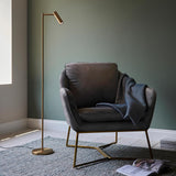 Endon Brackbourne floor lamp in brass with adjustable reader head, set beside a grey velvet armchair with throw against a green wall.