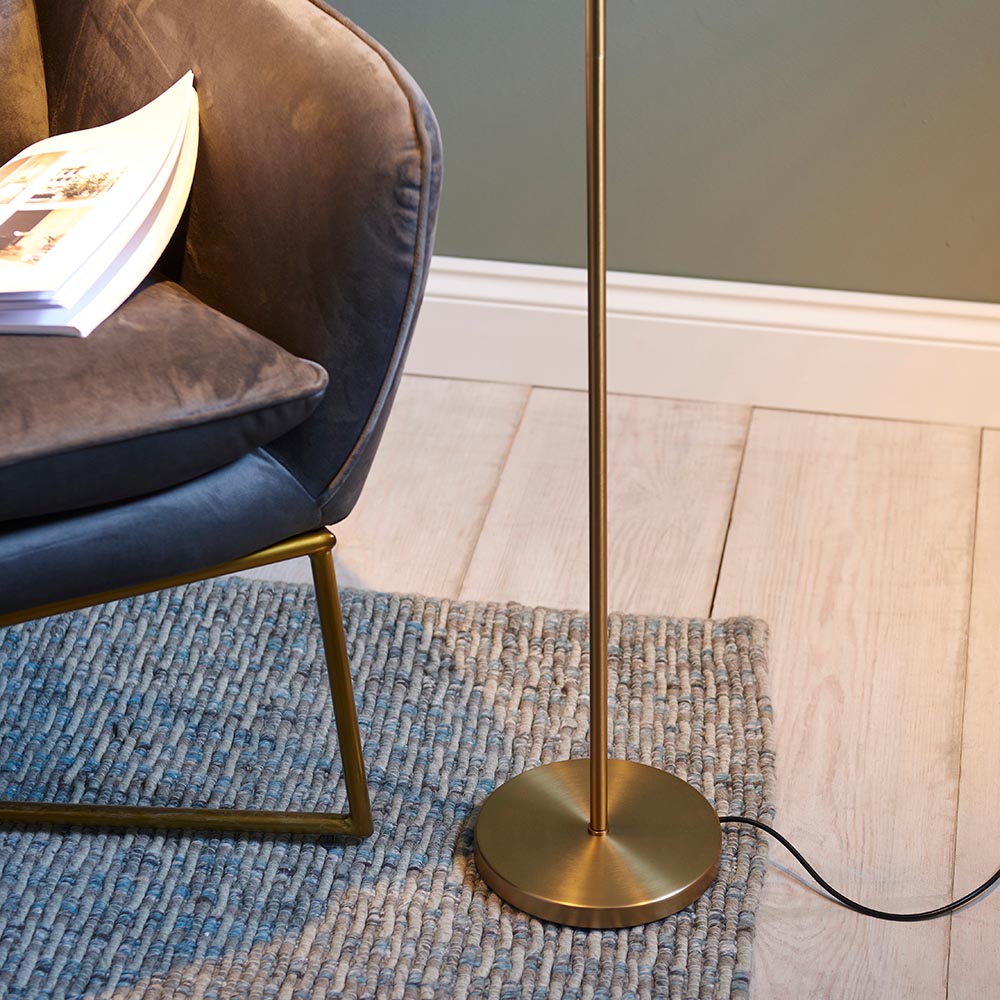Endon Brackbourne floor lamp base in brass finish beside a grey velvet chair on a woven rug, with black flex trailing across pale wood flooring.
