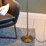 Endon Brackbourne floor lamp base in brass finish beside a grey velvet chair on a woven rug, with black flex trailing across pale wood flooring.