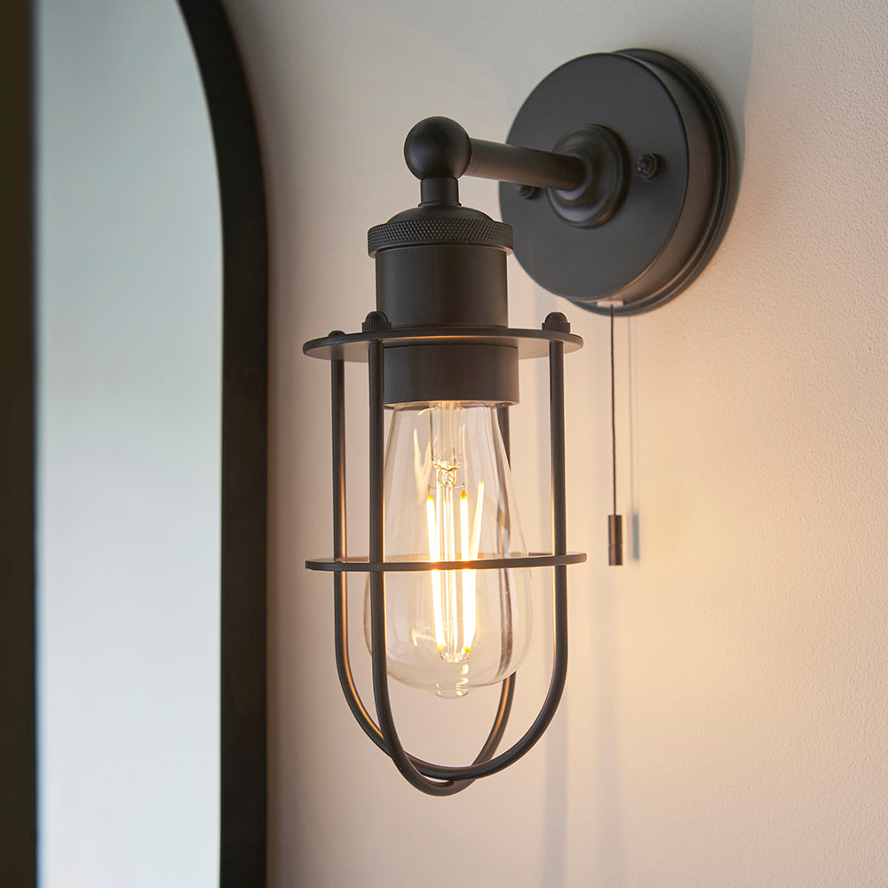 Bevwood industrial wall light in black metal with cage shade, E27 filament bulb and pull cord, shown mounted on a wall beside a mirror.