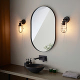 Bevwood black bathroom wall lights flank an oval mirror above a vanity, featuring industrial cage design, filament bulb, and pull cord.