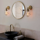 Bevwood industrial brass wall lights with cage design and pull cord flank a round mirror above a bathroom vanity with black basin.