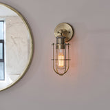 Bevwood industrial-style brass wall light with open cage design and lit filament bulb, mounted beside a round mirror on a pink bathroom wall.