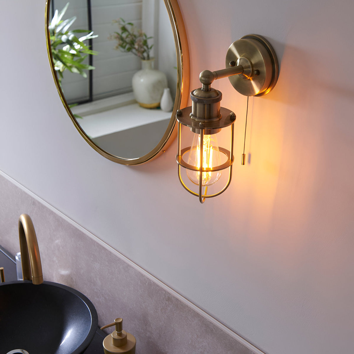 Bevwood Bathroom Wall Light in brass with industrial cage design and pull cord, mounted beside a round mirror above a black basin.