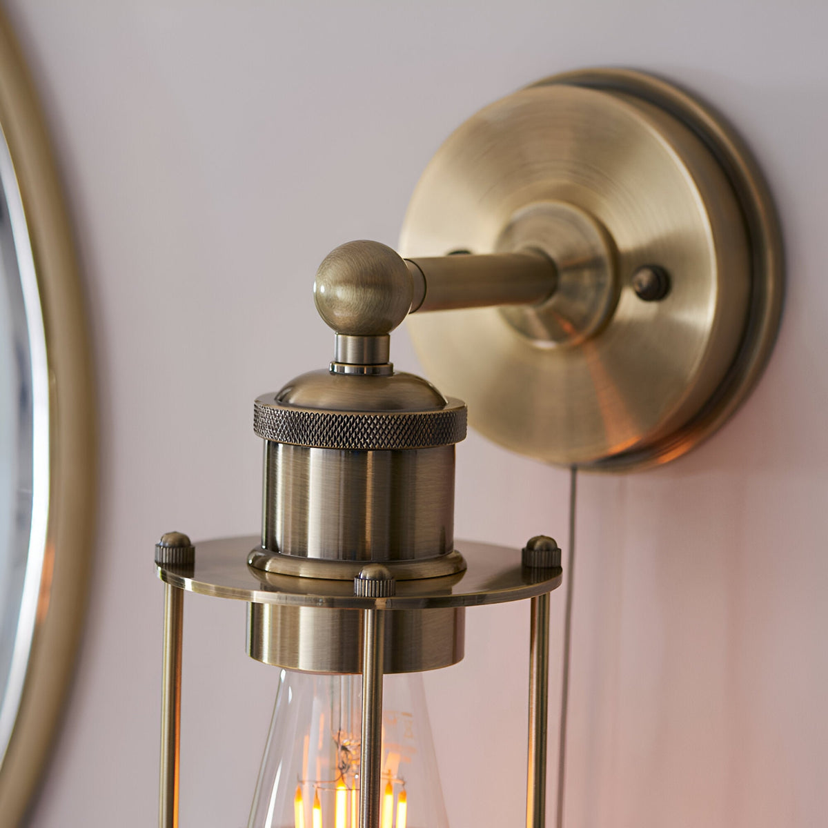 Bevwood industrial-style bathroom wall light in brass, close-up of knurled fitting, ball-jointed arm and round wall plate with visible E27 filament bulb.
