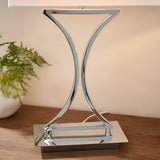 Endon Brackton table lamp base in chrome with curved hourglass frame on a rectangular plate, shown on a wood surface beside a fern in a bedroom setting.