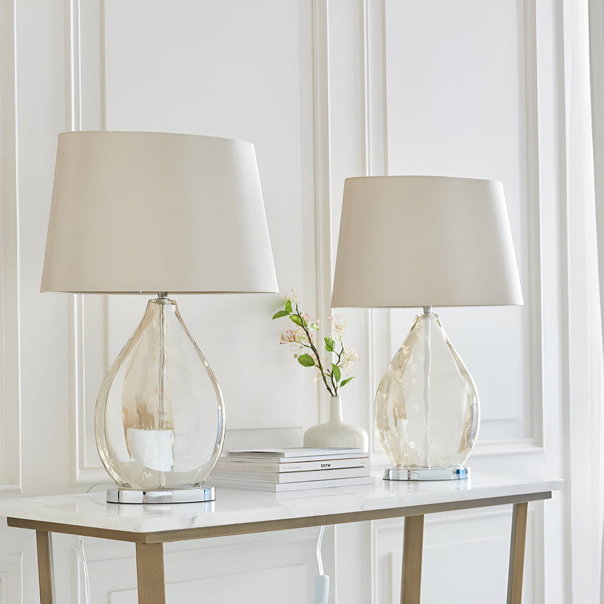 Brockstead Table Lamp Twin Pack in Gold, showing two teardrop glass lamps with chrome bases and mink oval shades, styled on a marble console table.