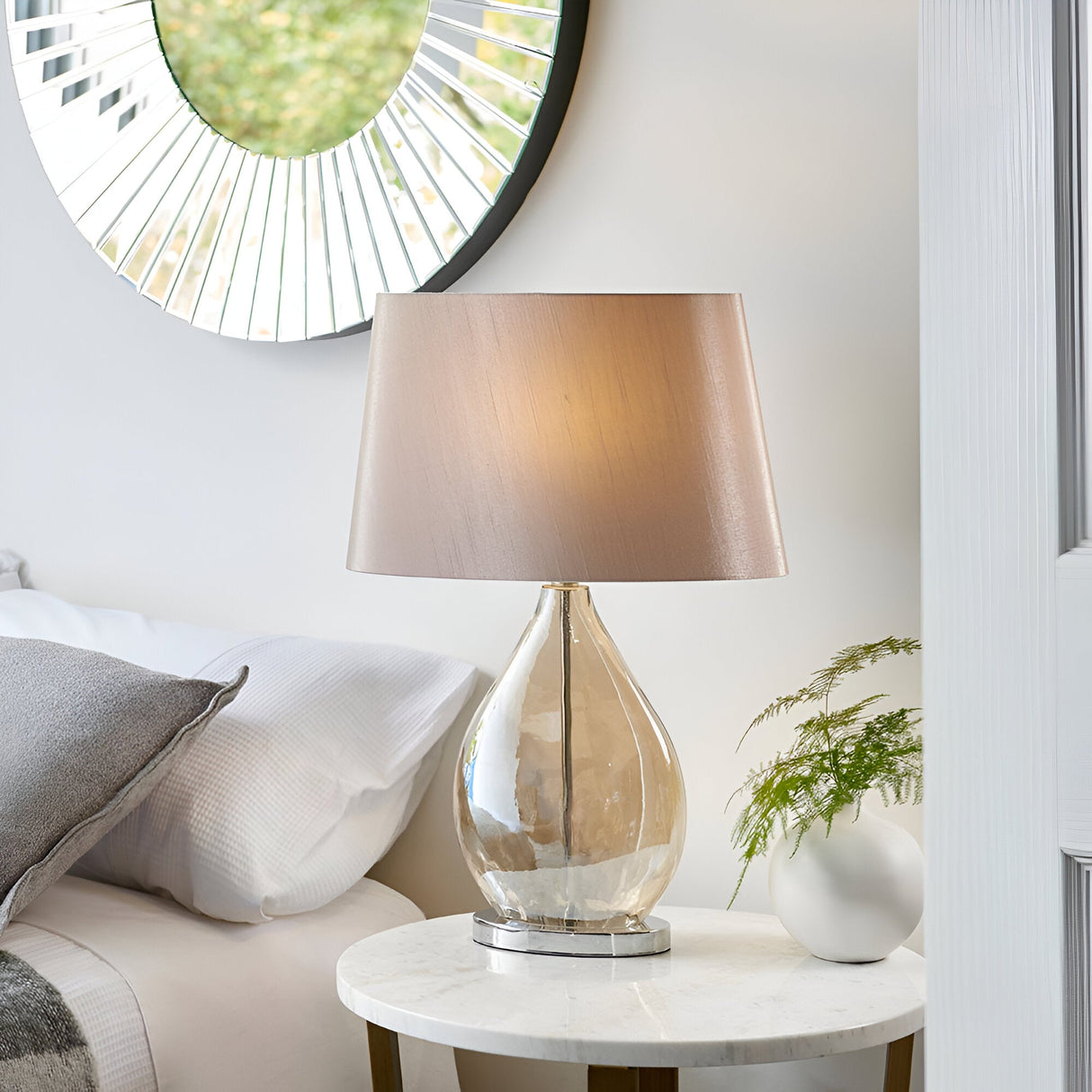 Brockstead Table Lamp in a bedroom setting, with a gold dimpled glass teardrop base, chrome fittings, and a mink oval faux silk shade on a marble side table.