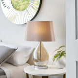 Brockstead Table Lamp in a bedroom setting, with a gold dimpled glass teardrop base, chrome fittings, and a mink oval faux silk shade on a marble side table.