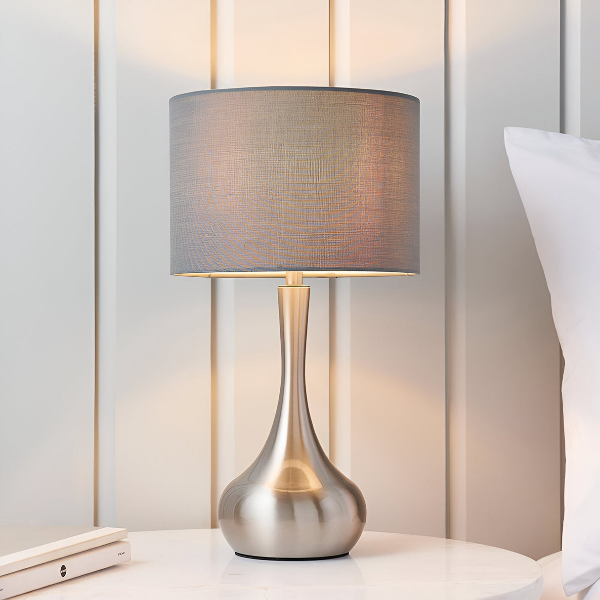 Endon Chartbury LED table lamp with satin nickel tulip-shaped base and grey fabric drum shade, shown on a bedside table beside a pillow.