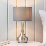 Endon Chartbury LED table lamp with satin nickel tulip-shaped base and grey fabric drum shade, shown on a bedside table beside a pillow.