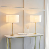 Endon Caldley twin pack table lamps with clear glass cylinder bases, brass fittings and white drum shades, lit on a marble console table.