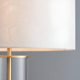 Close-up of the Endon Caldley table lamp showing its white faux silk drum shade, brass fitting and clear cylindrical glass base against a grey backdrop.