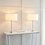 Endon Caldley twin pack table lamps with clear cylindrical glass bases, nickel fittings and white drum shades, lit on a marble console table.