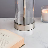 Close-up of the Endon Caldley table lamp's nickel base and clear cylindrical glass on a marble surface beside an open book and lit candle.