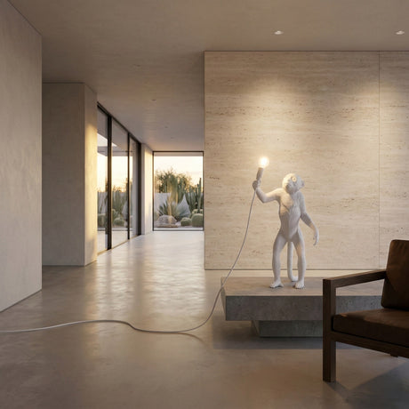A modern minimalist room with large windows overlooks outdoor plants. The Monkey Standing Table Lamp - White stands lit on a low platform next to a brown cushioned chair.