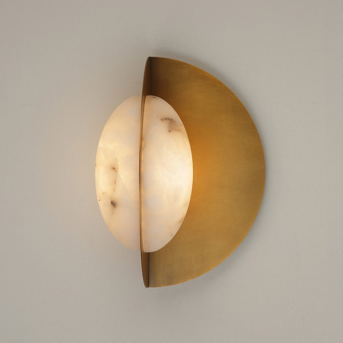 Althia LED wall light with a rounded alabaster diffuser set against a brushed brass half-disc backplate, warm white glow, shown on a plain light grey wall.