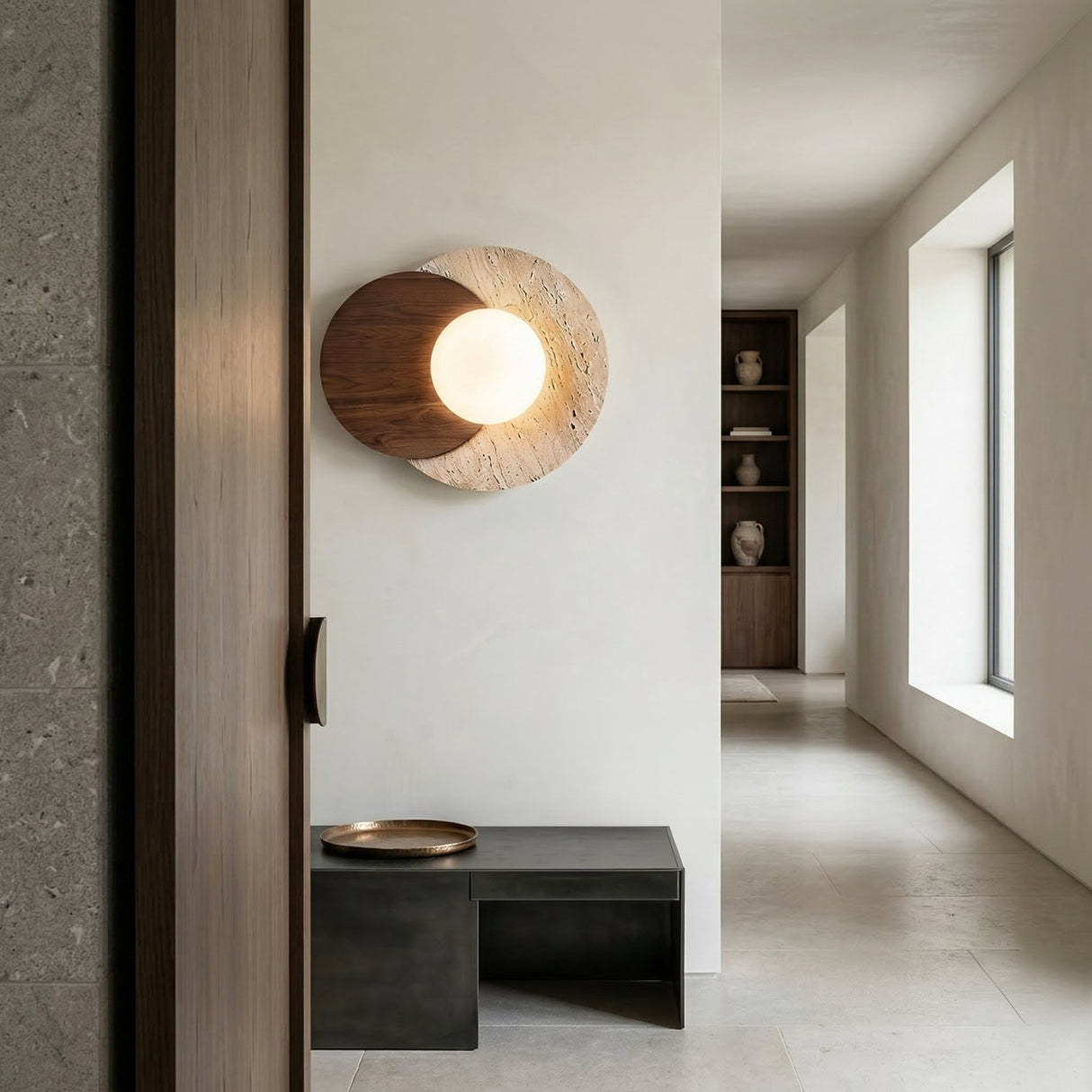 Minimalist hallway featuring the Zorella 1 Light Disc Travertine Wall Light in walnut brown & light beige, with wooden and stone details, a black bench with tray, light stone floor, white walls, shelves with pottery, and large windows.