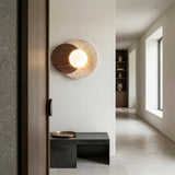 Minimalist hallway featuring the Zorella 1 Light Disc Travertine Wall Light in walnut brown & light beige, with wooden and stone details, a black bench with tray, light stone floor, white walls, shelves with pottery, and large windows.