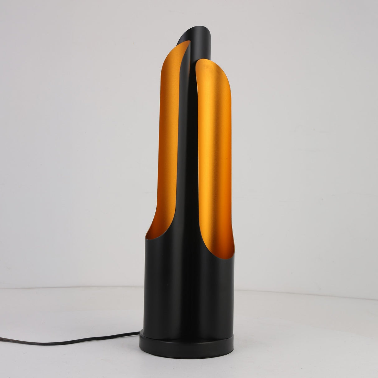 The Stealth Table Lamp - Brass & Black showcases a modern design with a cylindrical black base and two curved brass panels, creating a sculptural, flame-inspired look. It is plugged in and displayed against a plain white background.
