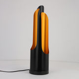 The Stealth Table Lamp - Brass & Black showcases a modern design with a cylindrical black base and two curved brass panels, creating a sculptural, flame-inspired look. It is plugged in and displayed against a plain white background.