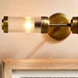 Akurdi 2 Light Bathroom Wall Light in Clear, ribbed glass cylinder with brass caps and collar on a wall-mounted arm, shown lit above a wooden-framed mirror.