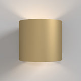 The Rond Up & Down Wall Light in matte gold, with its modern cylindrical shape, is mounted on a plain white wall to provide dual-directional lighting that softly glows both upward and downward.