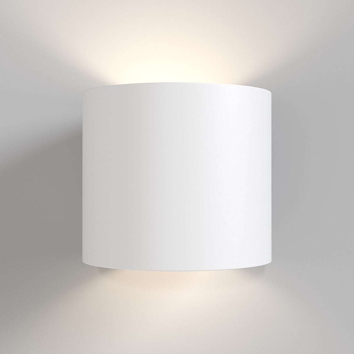 The Rond Up & Down Wall Light - White features a sleek, cylindrical design mounted on a light wall. Its minimalist style emits soft light from both ends, creating an ambient glow. The matte white finish lends elegance to any space.